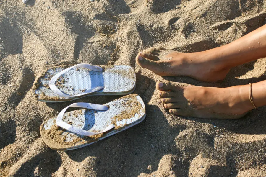 Women's feet in the sand beside thick blue flip flops.