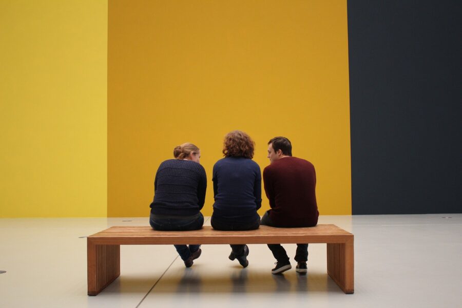 3 people sitting on a wooden bench at a contemporary art museum, with the walls painted bright yellow, mustard yellow, and navy blue