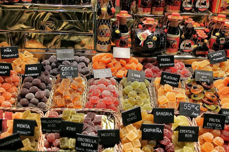 A display of various colorful food products and souvenirs in La Boqueria - a market with the best things to buy in Barcelona - including various flavors of sweets and bottles filled with a dark brown substance