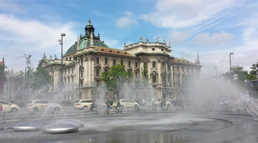 The spray of the fountain waters at Karlsplatz-Stachus, the first stop on our free walking tour of Munich, with views of white cars driving by and the architecture of a stone building on a bright sunny day