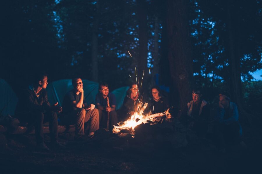 A group of seven friends surrounding a sparking campfire in the late evening