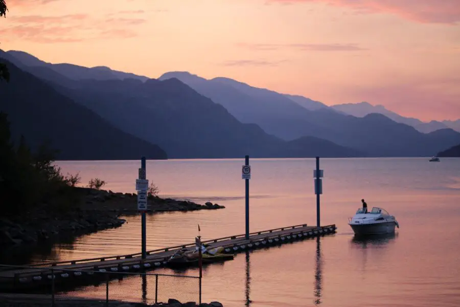 A man in a white speed boat beside a long wooden empty dock on Harrison Lake at sunset, the sky pink, purple, and orange
