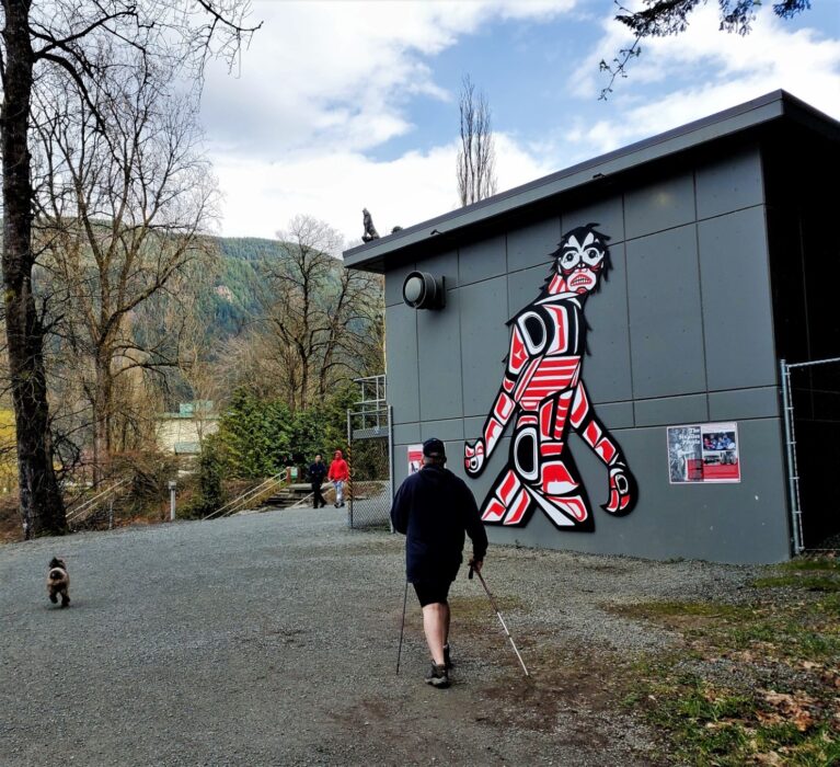 A man with hiking poles and a dog walking by the Sasquatch art piece on one of the flat and easy Harrison Hot Springs hikes