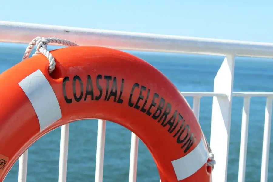 A bright orange ring buoy with "Coastal Celebration" written on it on the white rails of a BC ferry, surrounded by blue water