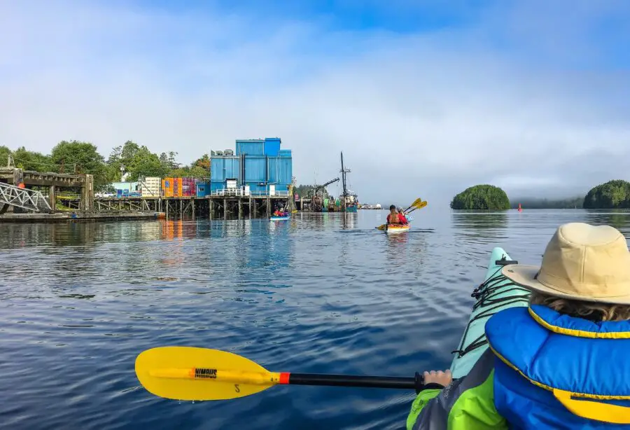 A few people in kayaks off the coast of Ucluelet BC, paddling by some brown docks and colorful buildings