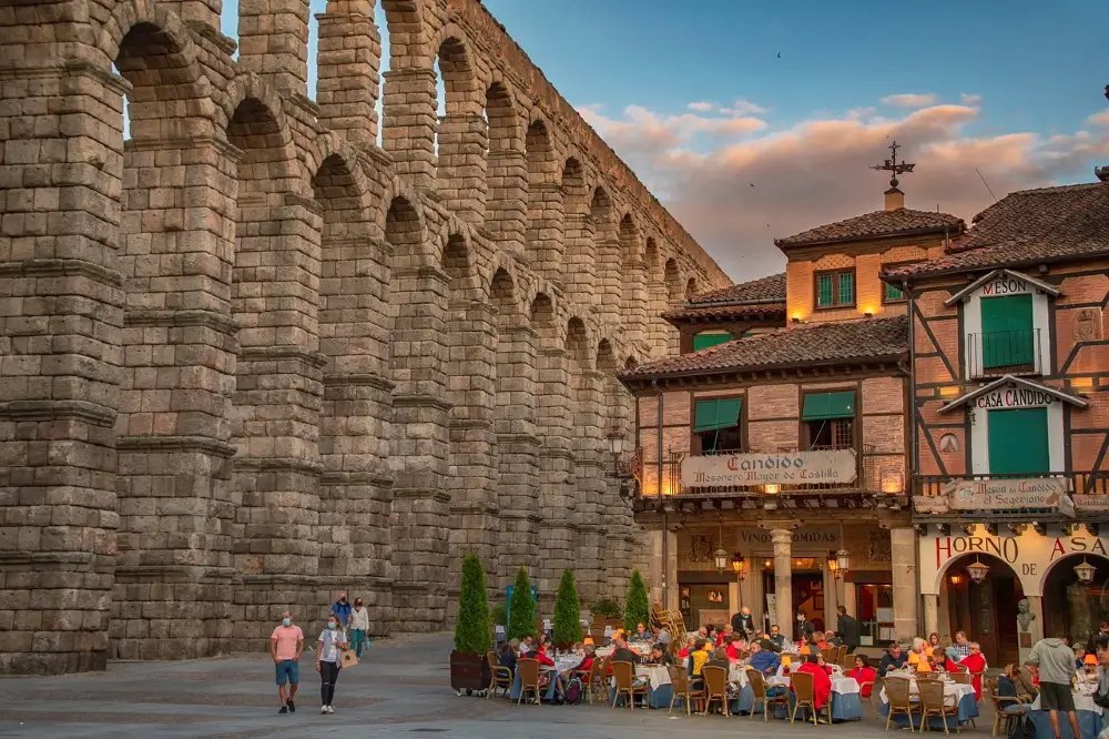 Photo of the Segovia restaurants next to the Aqueduct at dusk, with several people eating outside in the evening light.