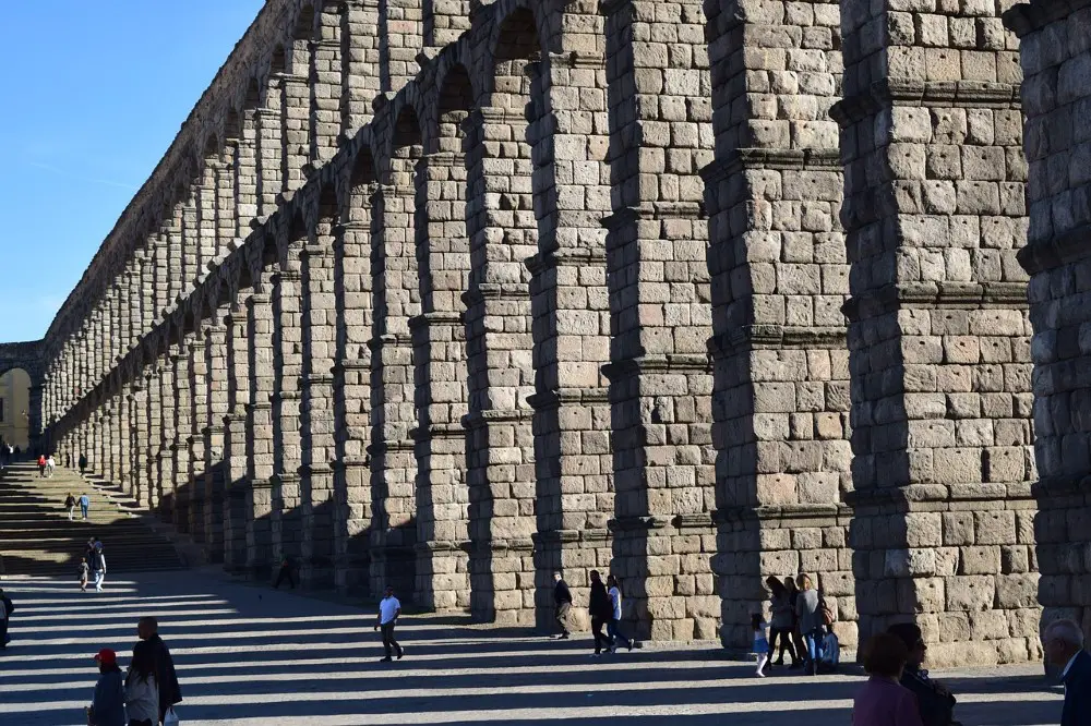 Photo from the base of the Aqueduct Segovia on a sunny day, with several visitors walking in its shadows.