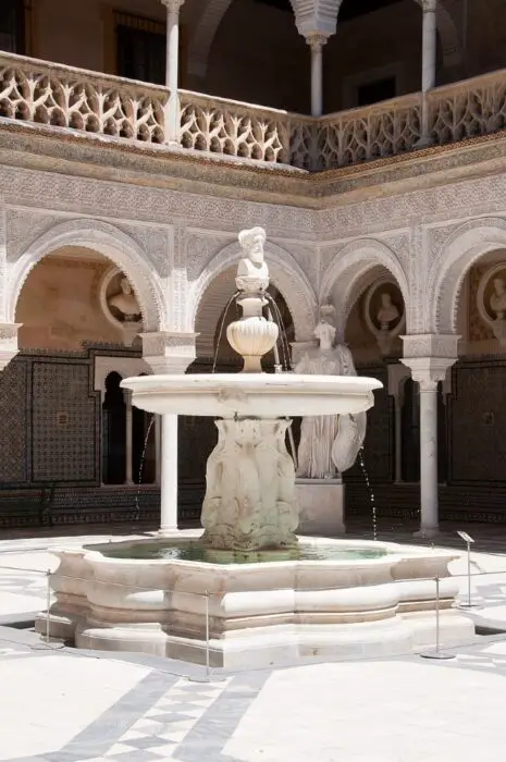 A white fountain in the middle of the Casa de Pilatos square bathed in sunlight.