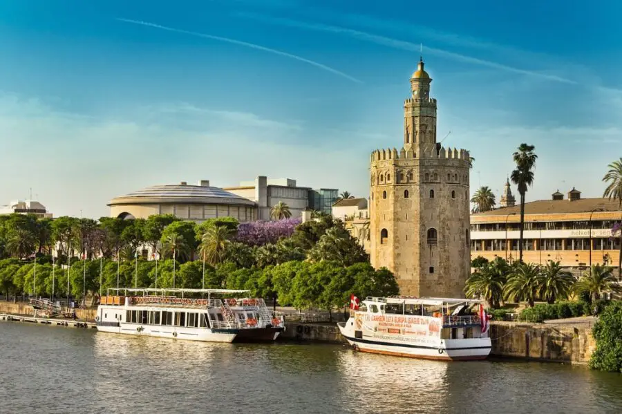 A view of the Torre del Oro basking in the sunlight with two white boats parked on the riverside, one of the best walks in Seville.